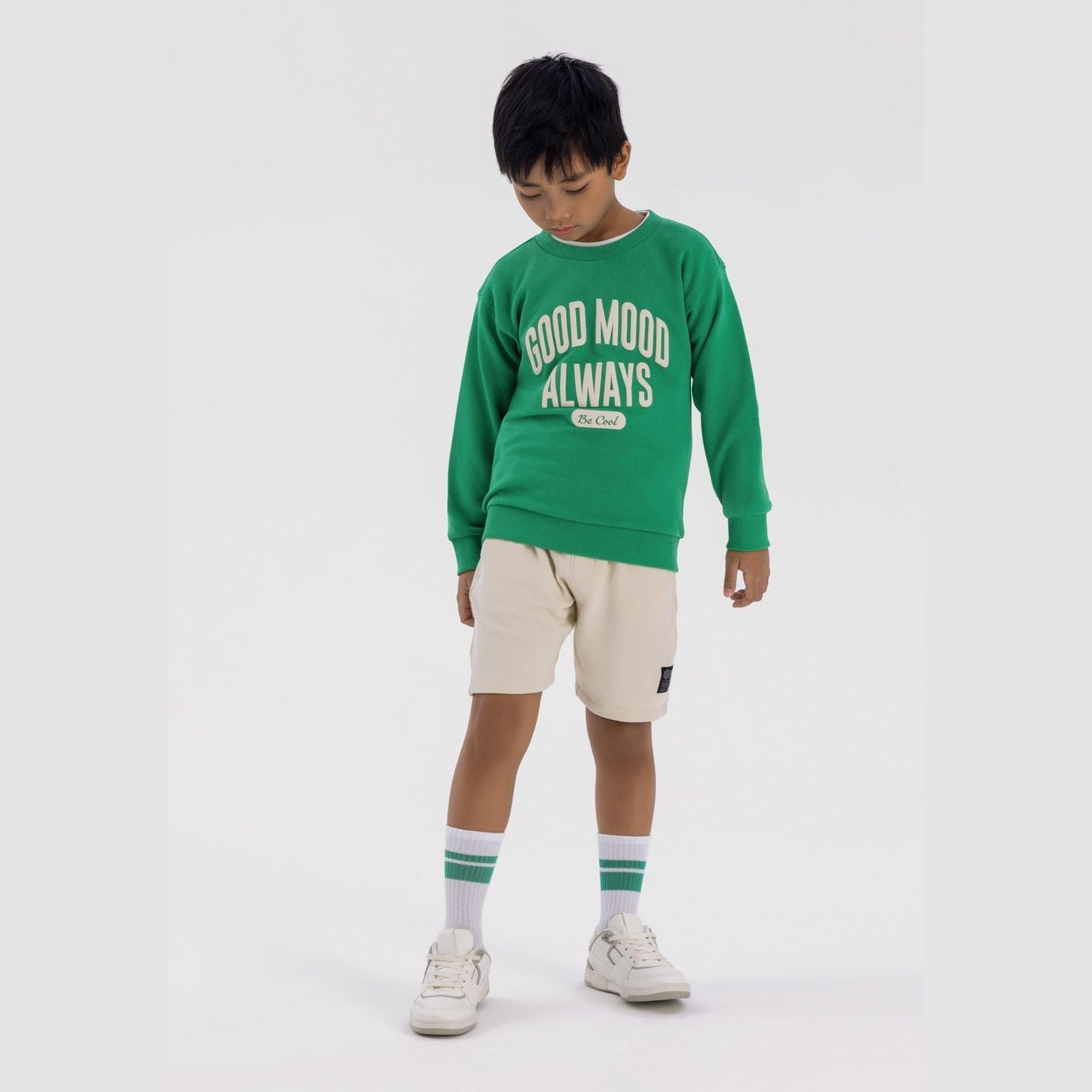 Boy wearing bright green oversized sweatshirt with Good Mood Always print, paired with beige shorts and green striped socks on white background