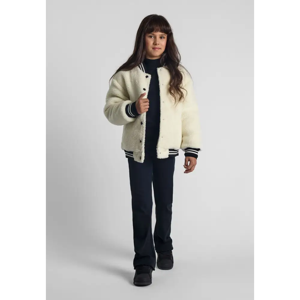 Young girl wearing cream borg baseball jacket with navy ribbed trim, paired with navy trousers and black boots, standing against white background