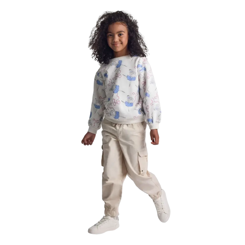 Young girl wearing grey cargo trousers with white floral print sweatshirt, standing against black background with one foot raised in playful pose