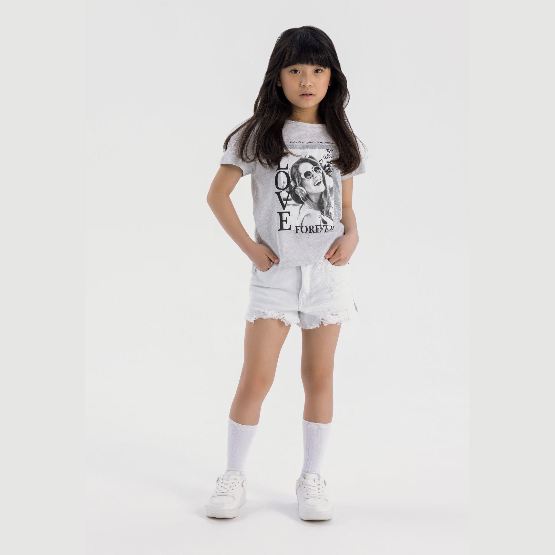 Young girl modeling white denim shorts with frayed hem and white graphic t-shirt, full body front view against light background