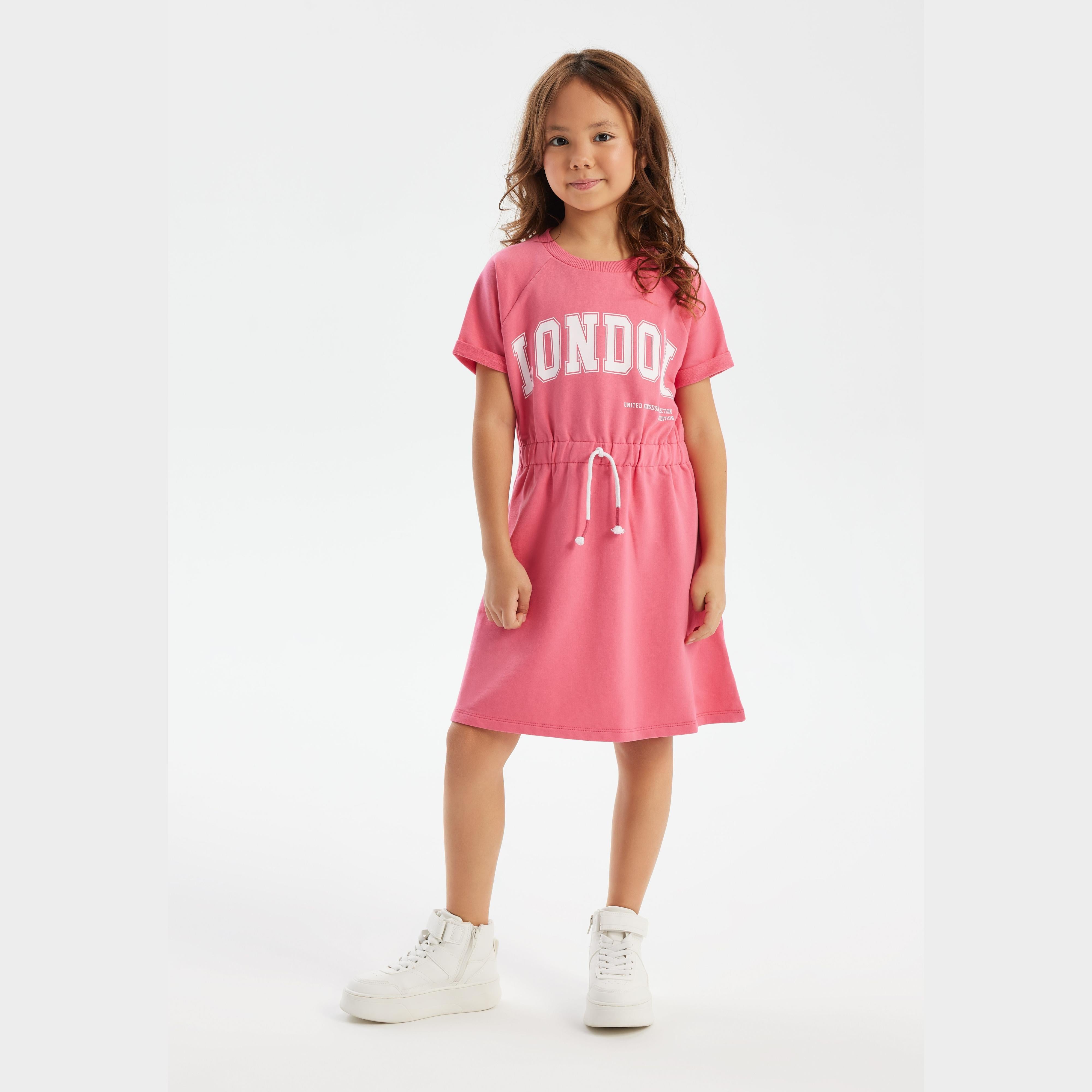 Young girl wearing pink fleece dress with London print, white sneakers, standing against white background