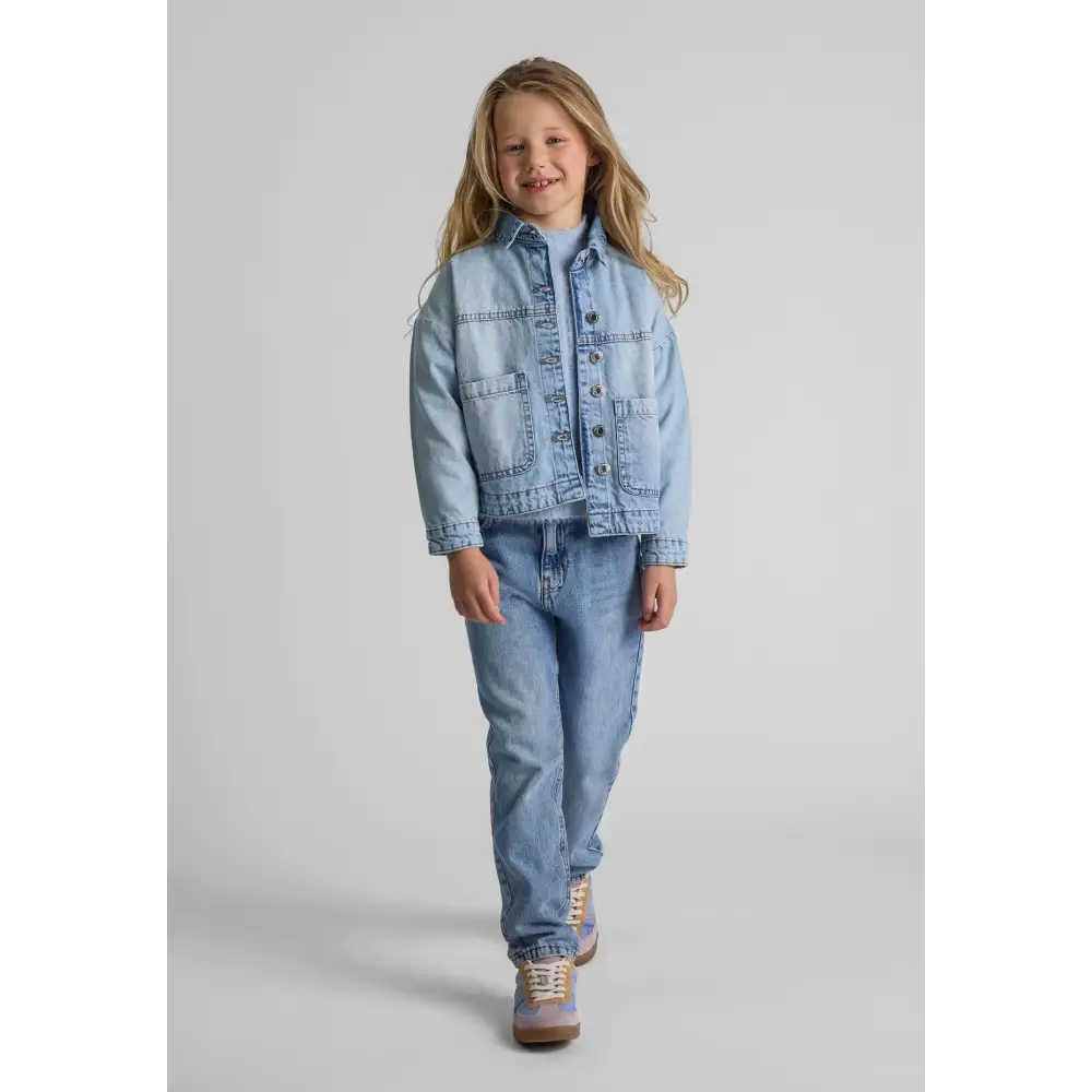 Young blonde girl wearing oversized light blue acid wash denim jacket with matching jeans, standing against white background with hands at sides