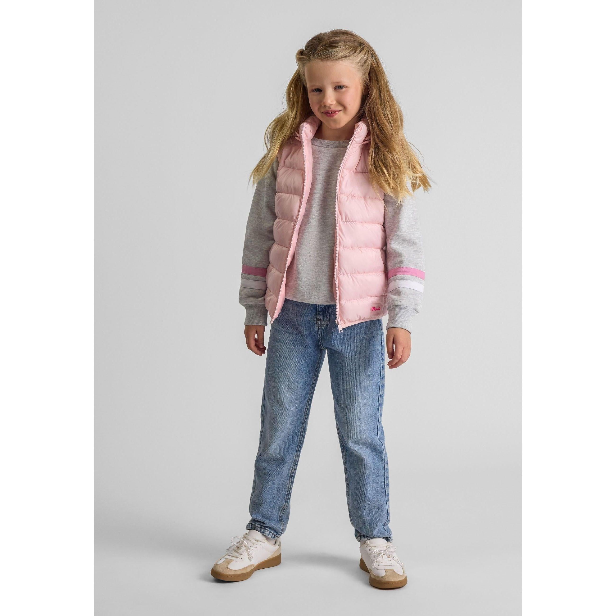 Young girl wearing pink quilted gilet over grey sweatshirt with blue jeans and white trainers, smiling against neutral background