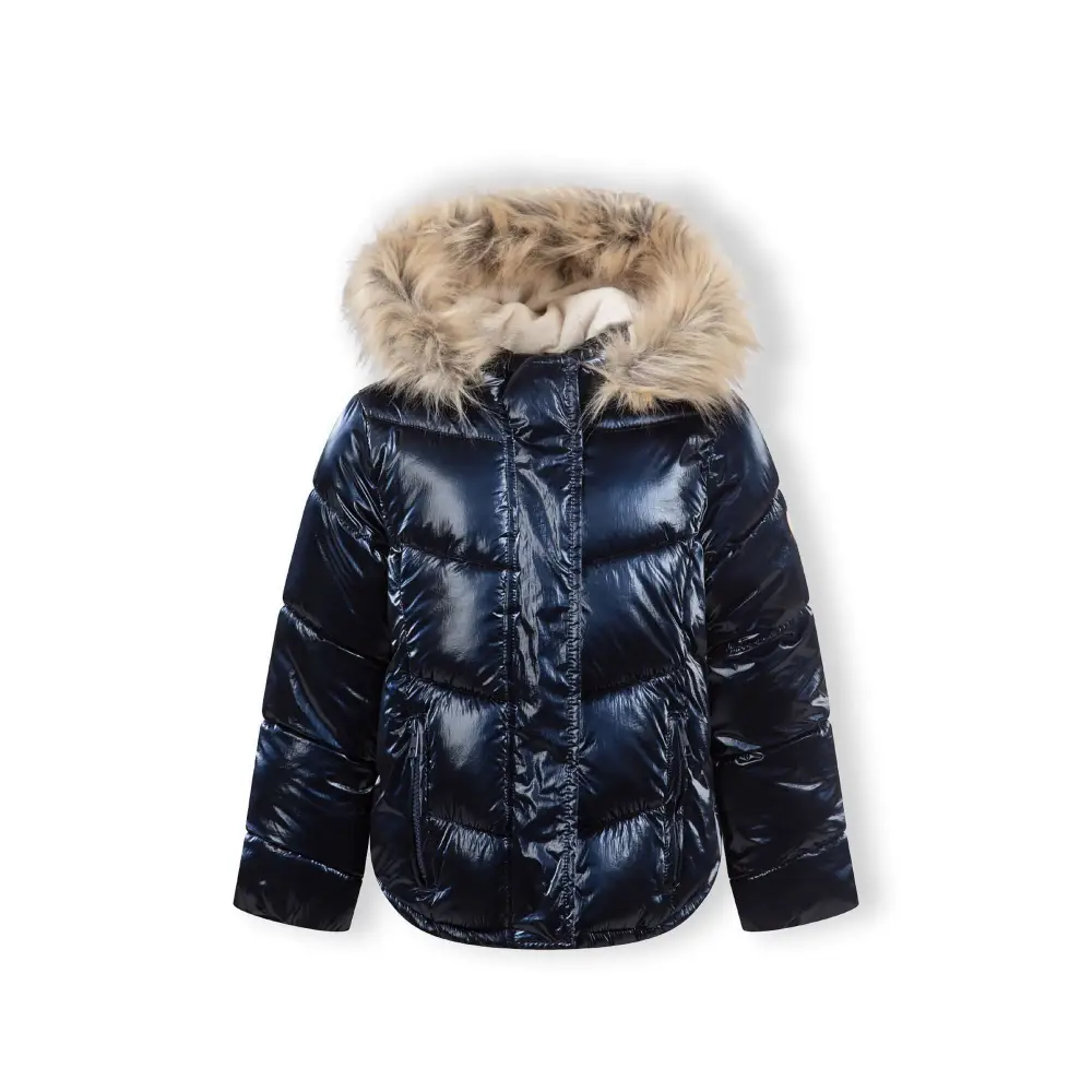 Young girl in navy metallic winter jacket with hood up showing faux fur trim, wearing light blue jeans and white shoes, side angle against white background