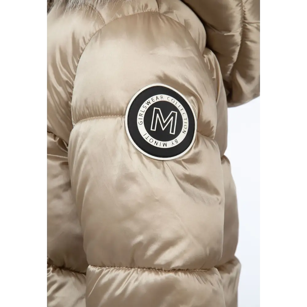 Close-up detail of gold quilted jacket showing Minoti circular logo patch on sleeve with white faux fur hood trim visible in background