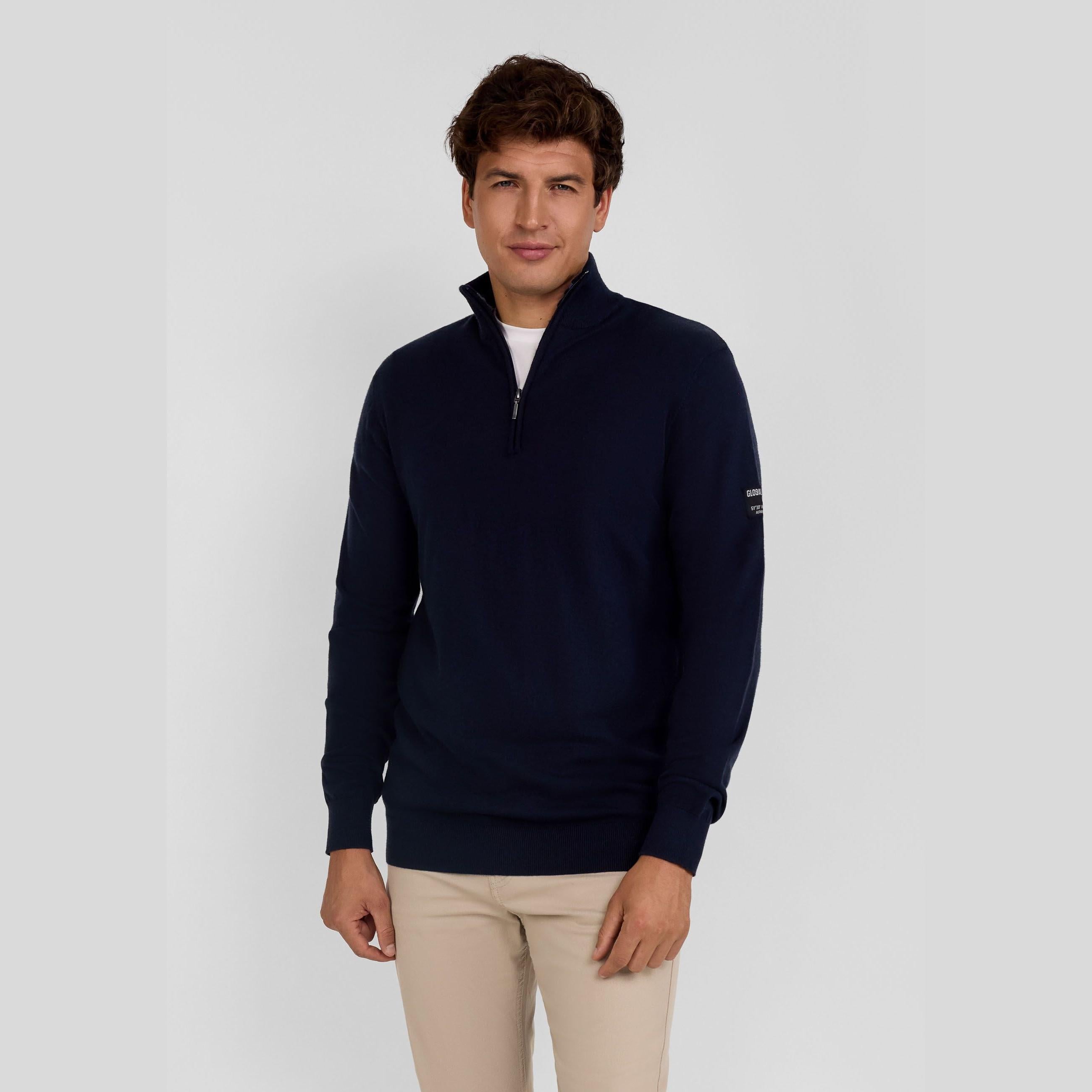 Male model wearing navy half-zip jumper with beige trousers against grey studio background, three-quarter view showing collar detail and fit