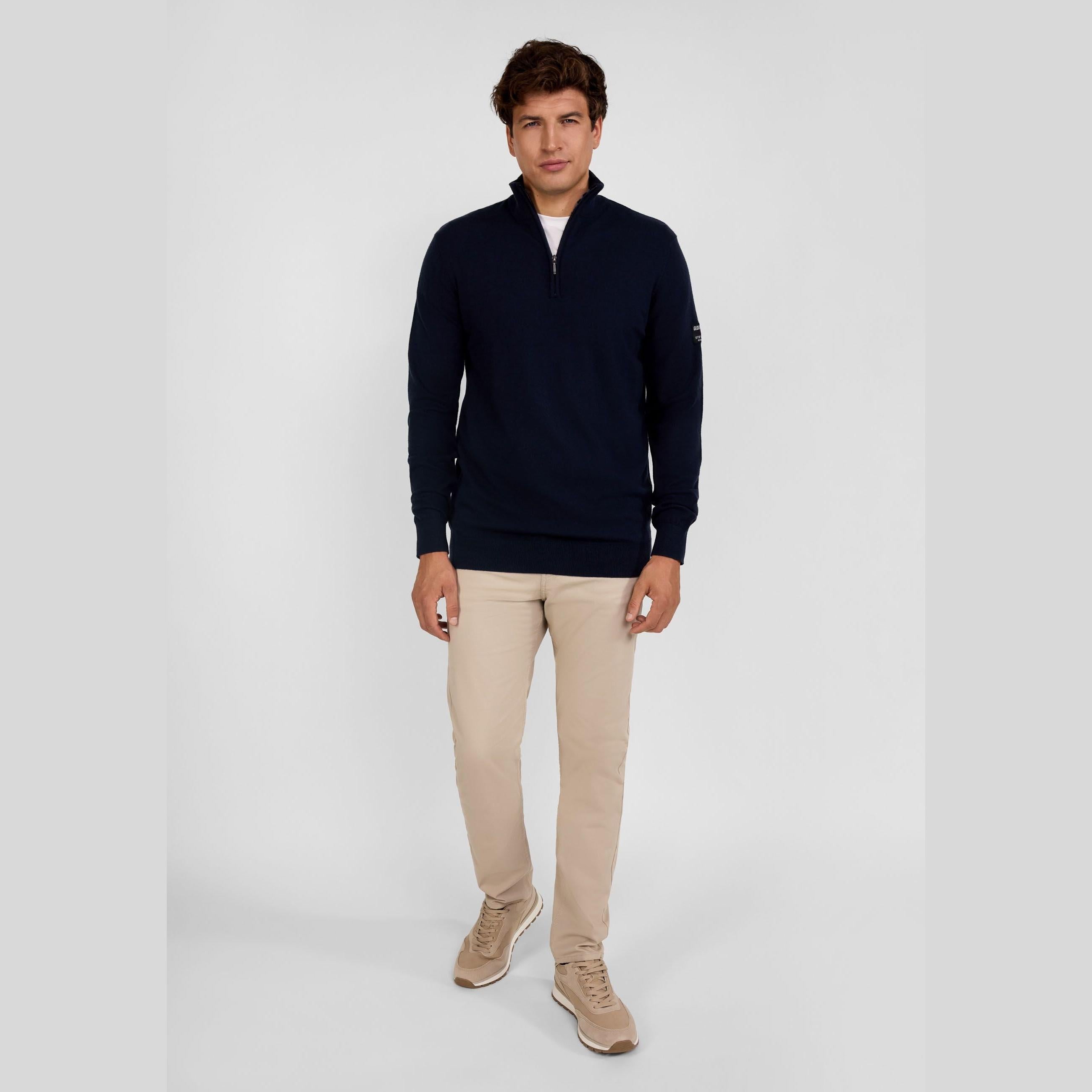 Full body shot of male model in navy half-zip jumper and beige chinos with beige sneakers against grey studio background